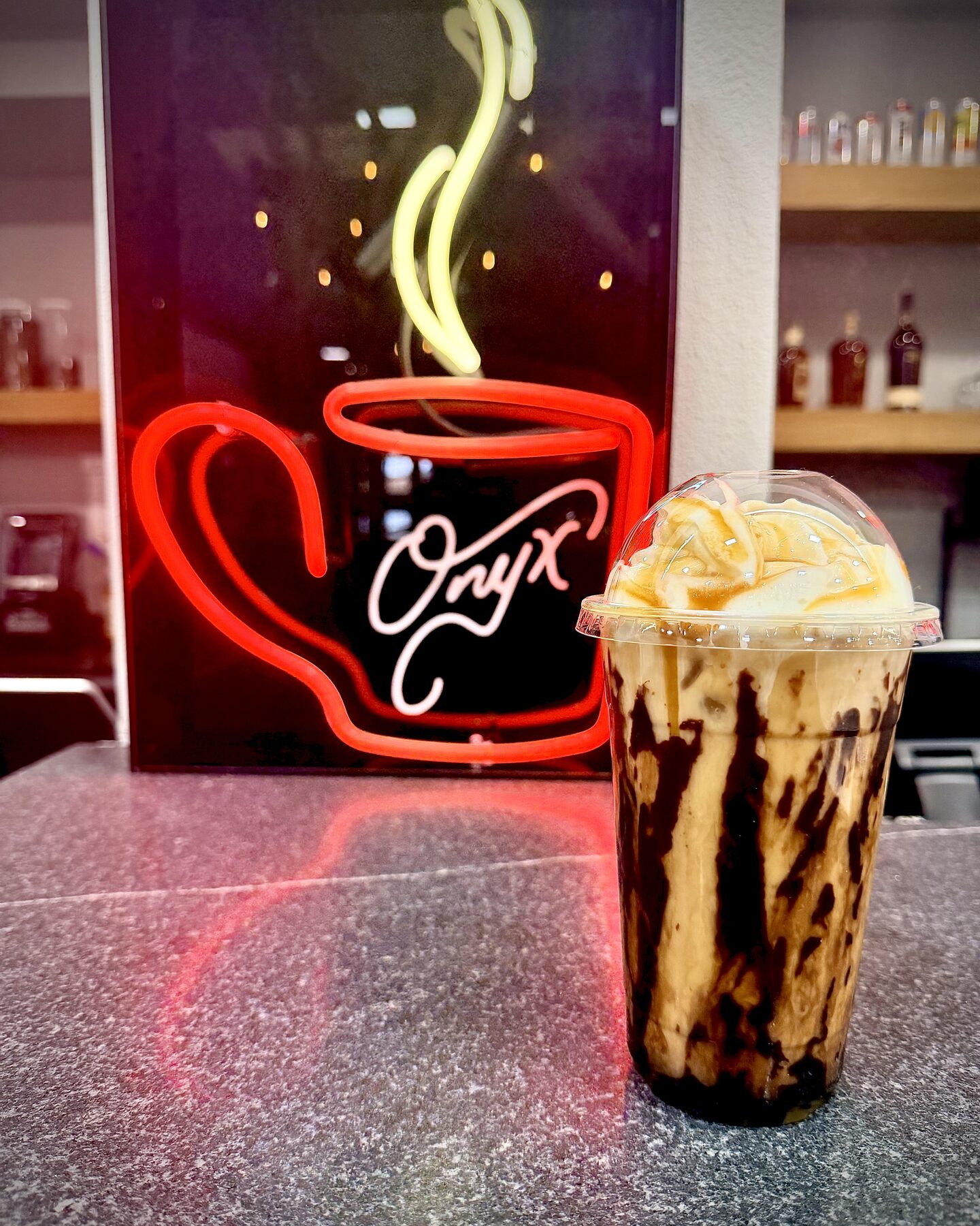 An iced coffee drink topped with whipped cream and caramel in front of the red neon Onyx coffee cup sign at the counter.