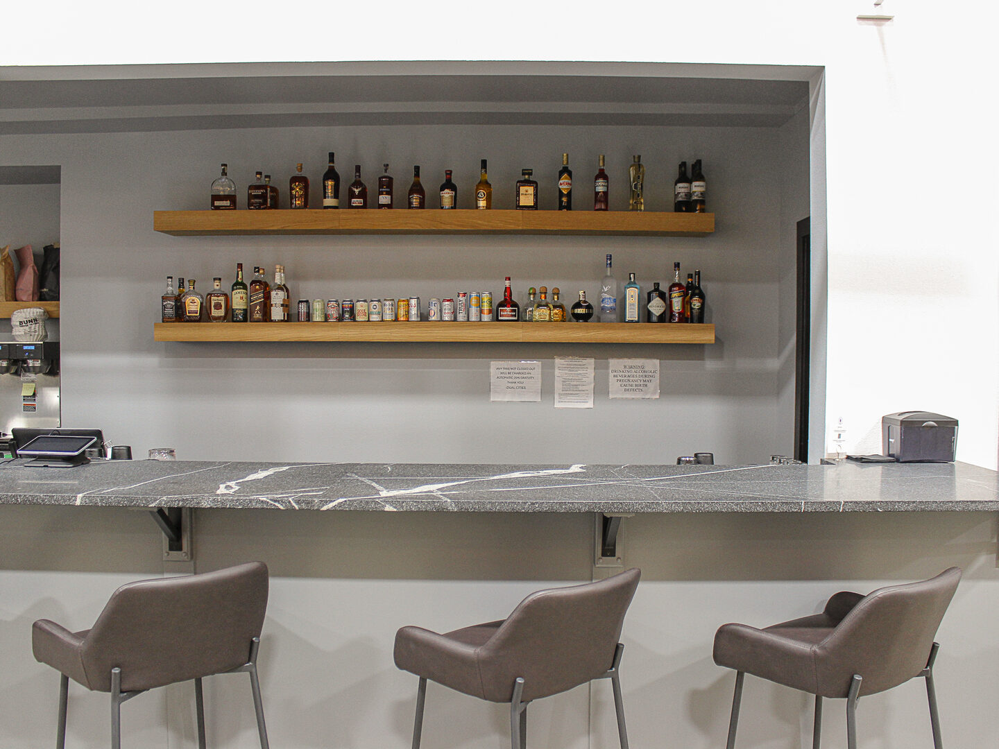 The bar at Dual Cities NWA, bar seating along the granite counter with shelves of bottles behind.
