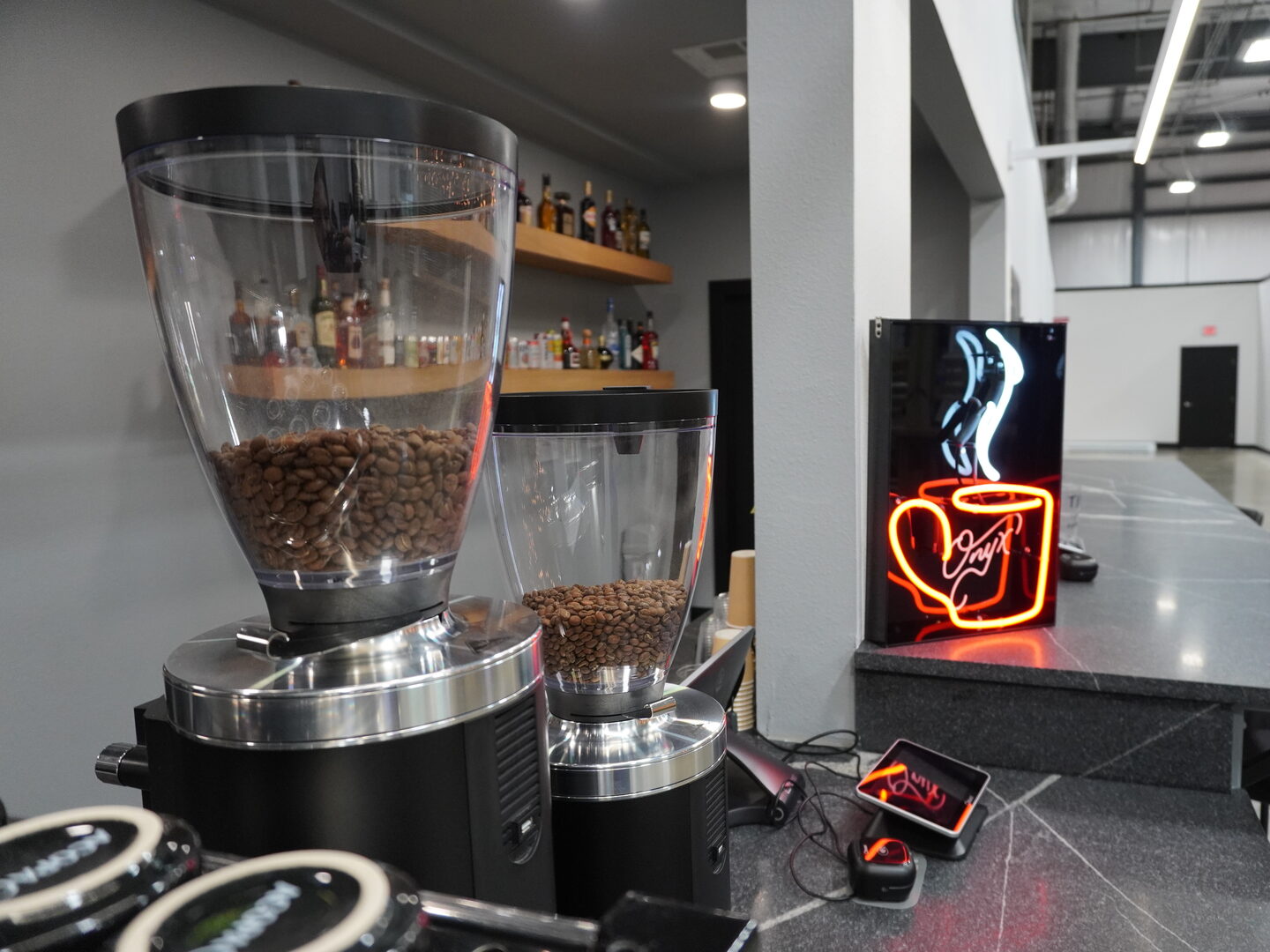 Two coffee grinders with hoppers full of whole beans on the dark stone counter, beside a red neon sign of a steaming Onyx coffee cup.