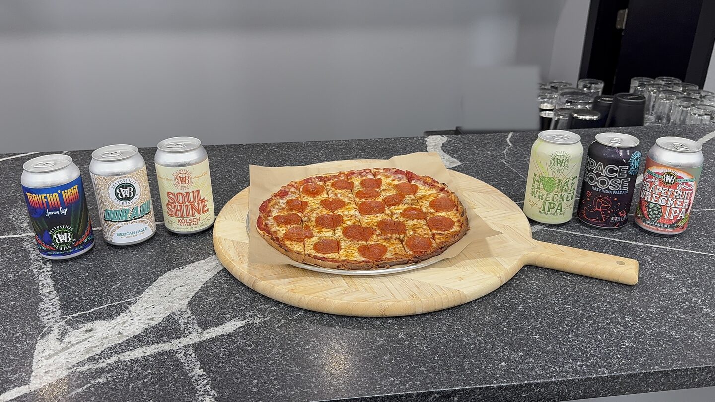 Tavern-cut pepperoni pizza on parchment on a round wooden serving board on the bar counter, cans of local craft beer on each side.