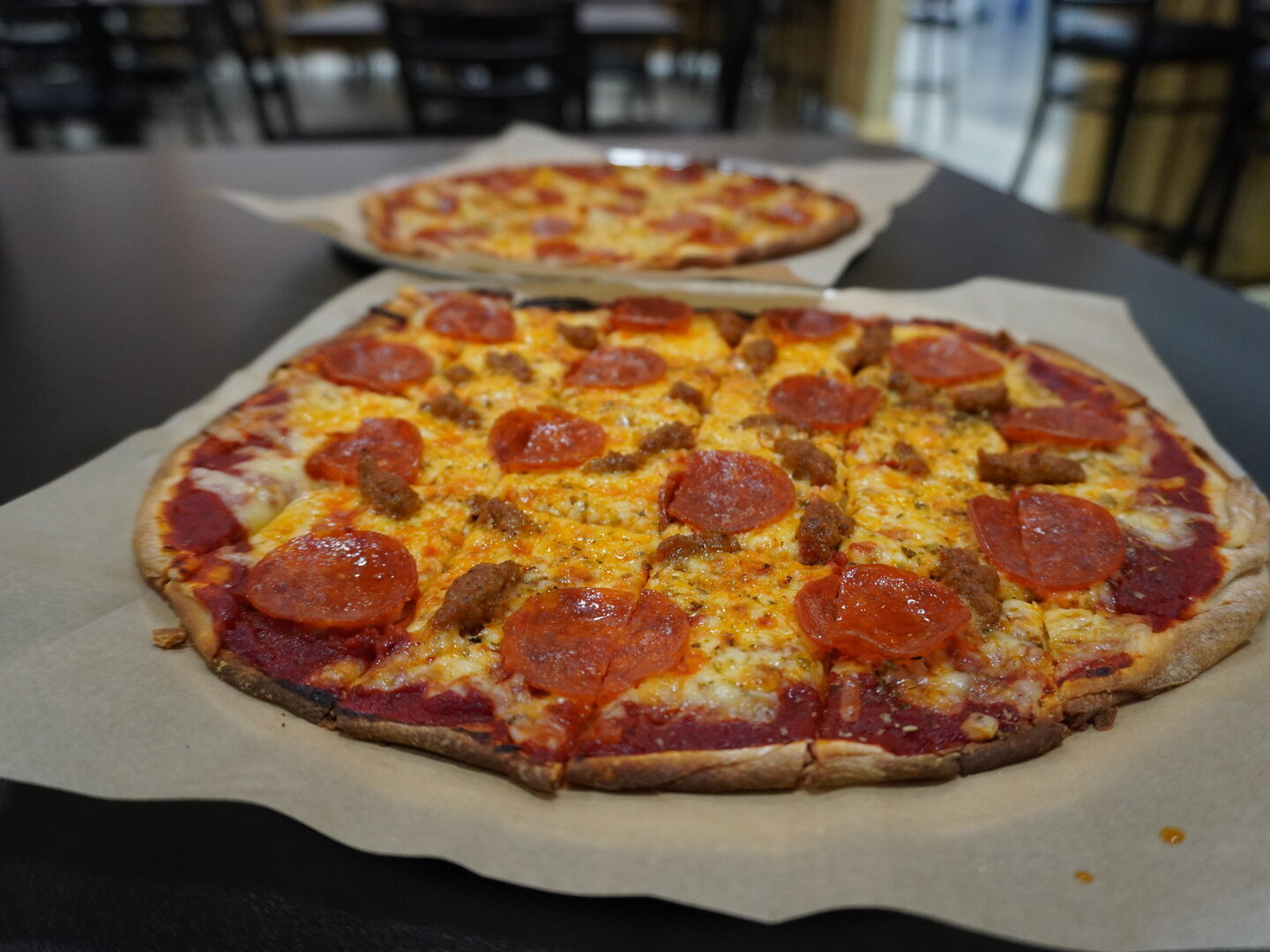 Two tavern-cut pizzas on parchment paper on a dark tabletop, the nearer one topped with pepperoni and sausage.