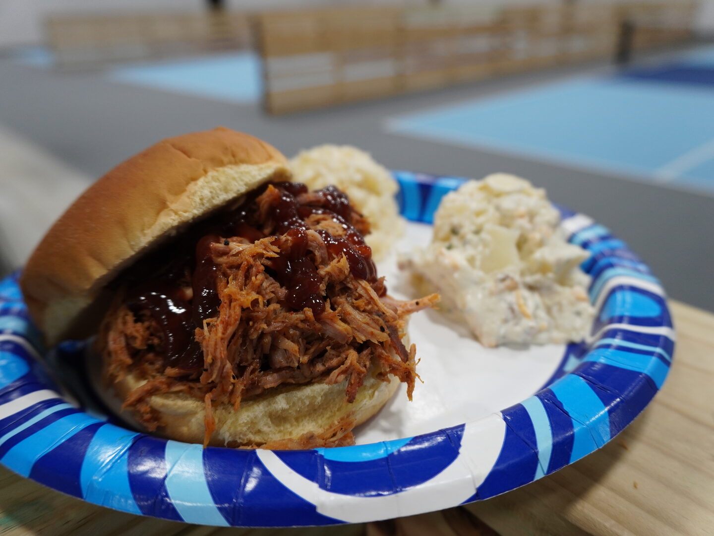 Pulled pork sandwich topped with barbecue sauce on a plate with potato salad and coleslaw, indoor pickleball courts in the background.
