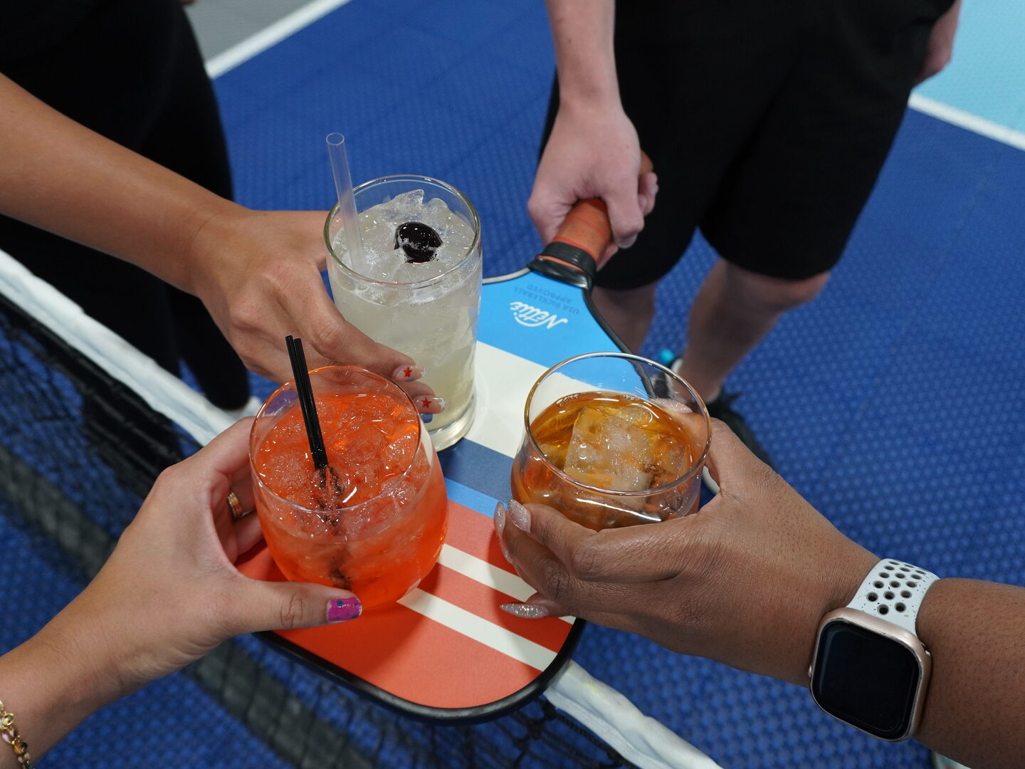 Three cocktails raised together over a pickleball paddle at the net on an indoor court.