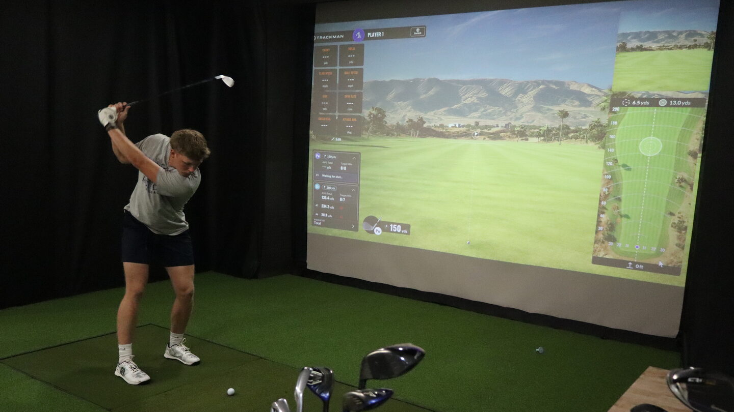A player at the top of his backswing in a golf bay at Dual Cities NWA, the course and shot data on the TrackMan screen.