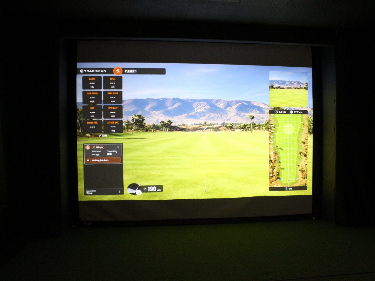 A Trackman golf simulator screen lit up in a dim bay, fairway on the display.