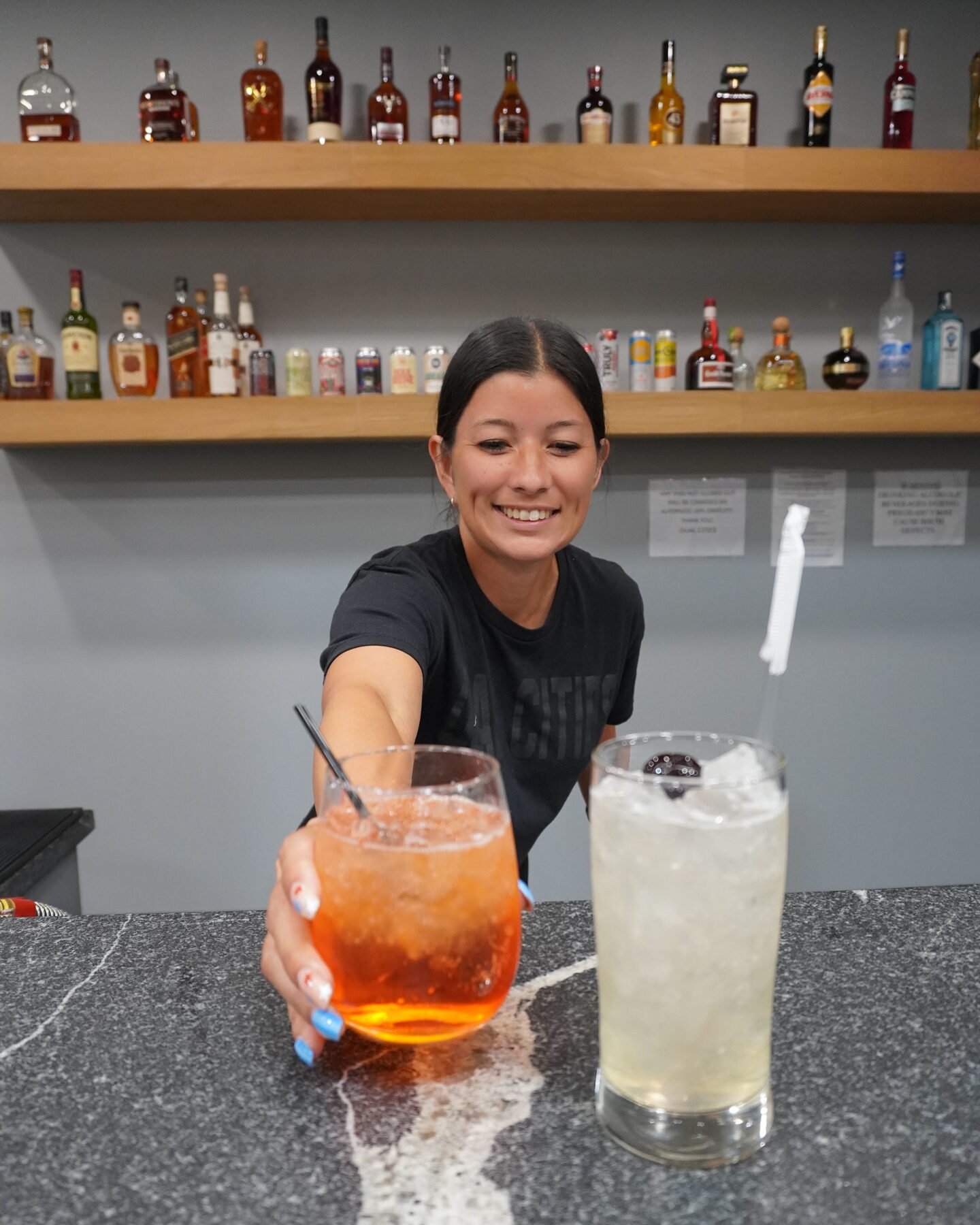 A smiling bartender reaches across the granite bar top to hand over an orange cocktail, shelves of bottles on the wall behind.