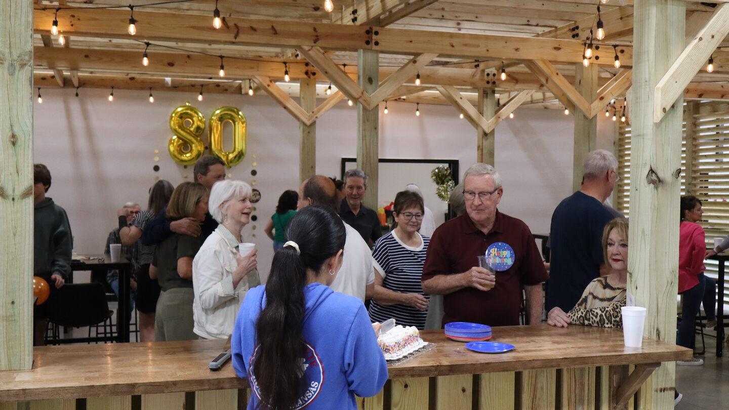 Guests mingle at an 80th birthday party under the wooden pergola strung with lights, gold 80 balloons on the wall and a birthday cake on the bar top.