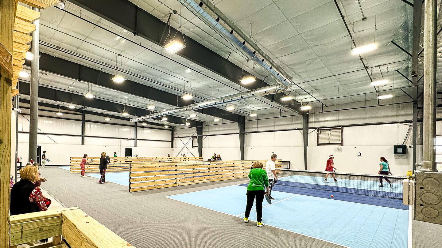 Open play on the indoor pickleball courts at Dual Cities NWA, players mid-game and friends watching from the rail.